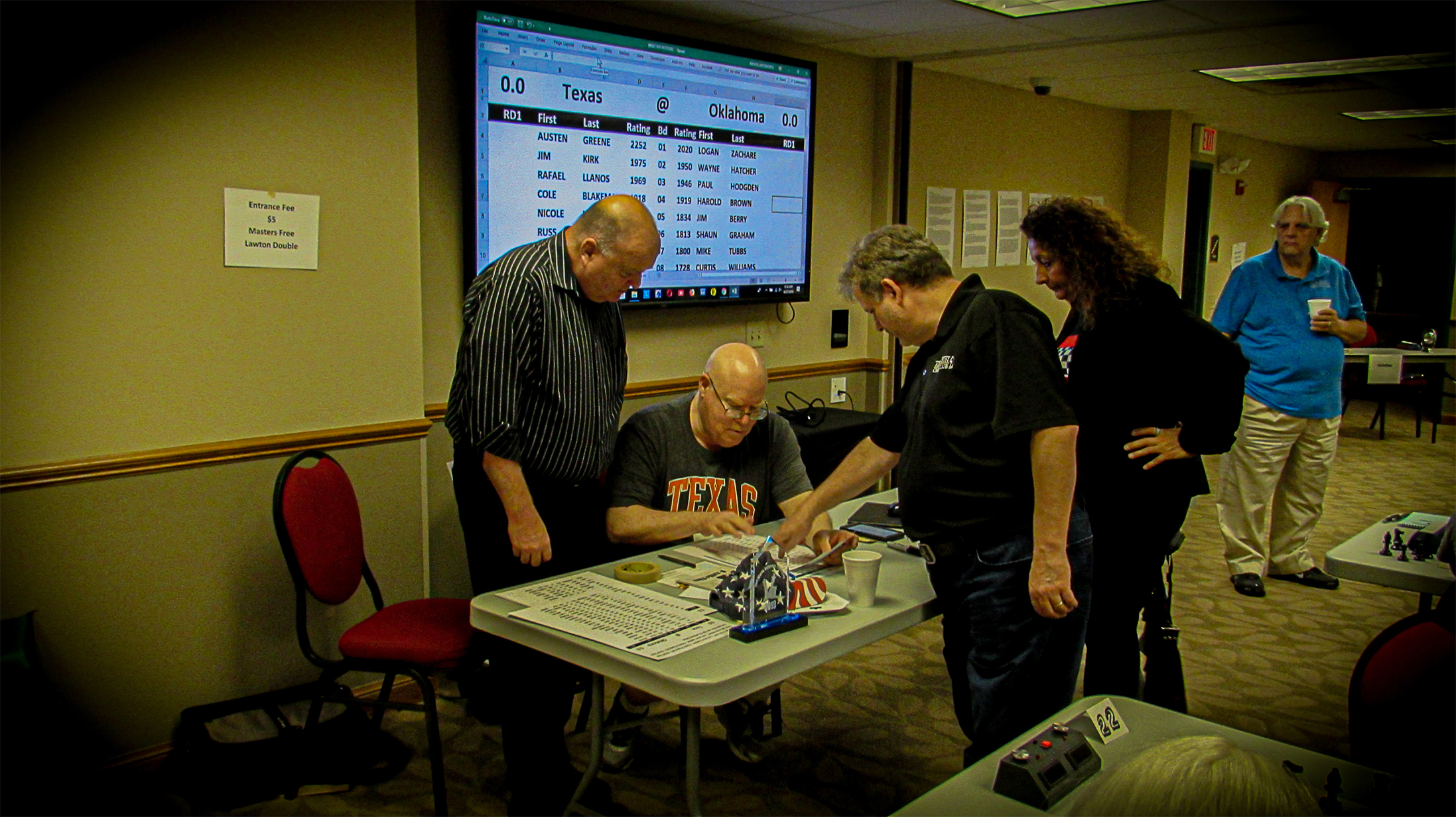 Both Team Captains huddle with Chief TD Jim Hollingsworth (second from left) to determine which Murray County Open players are available to fill two match section slots (one for each team).  Oklahoma Captain Phil Stegall (far left) ponders the "Jim Hollingsworth Solution" as Texas Captain Chris Wood (third from left) points out an adjustment for Boards 10 and 14 to better align the ratings.  Texan Sheryl McBroom (fourth from left) looks on.  In the background is Texan Troy Gillispie.  Photo by Mike Tubbs.